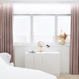 Wnętrze nowoczesnego salonu kosmetycznego z białym fotelem zabiegowym, białymi meblami, lustrem w złotej ramie i dekoracyjną tapetą w liście na ścianie, oświetlone naturalnym światłem z okna...