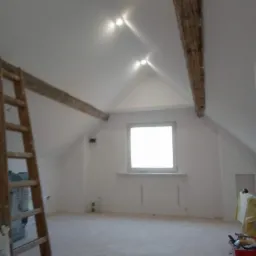 Poddasze w Lubinie po malowaniu, z widocznymi drewnianymi belkami stropowymi, nowymi punktowymi lampami sufitowymi, oknem i drabiną opartą o ścianę.