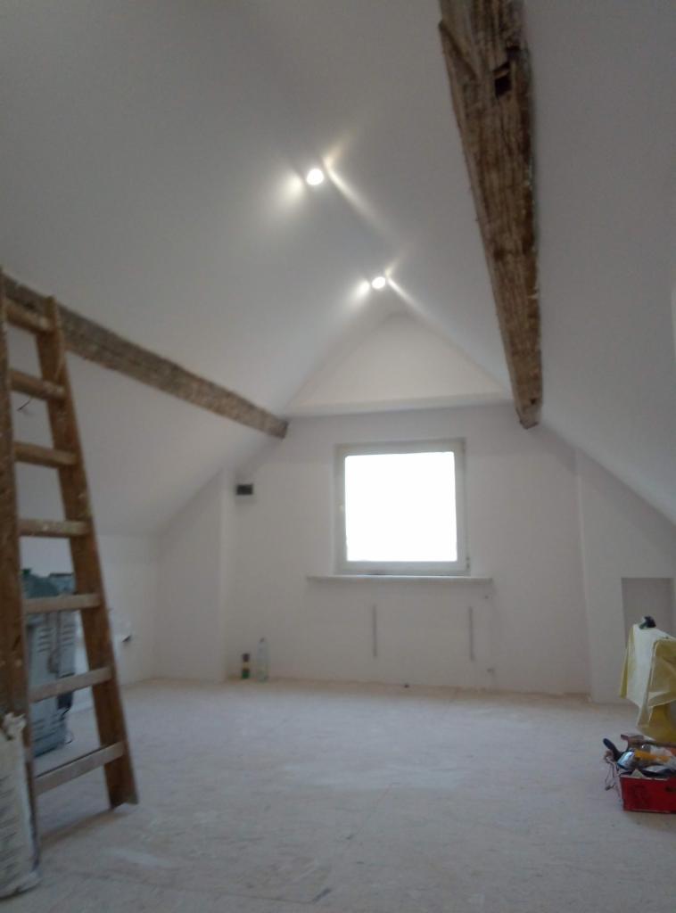 Poddasze w Lubinie po malowaniu, z widocznymi drewnianymi belkami stropowymi, nowymi punktowymi lampami sufitowymi, oknem i drabiną opartą o ścianę.