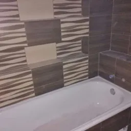 Wykończona łazienka z wanną, brązowo-beżowe płytki z motywem falistym na ścianie, widoczne wystające elementy obudowy wanny.