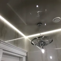 Błyszczący sufit z wbudowanym oświetleniem LED wzdłuż ścian i nowoczesną, okrągłą lampą wiszącą na łańcuchu; widoczny fragment białych drzwi z ozdobną ramą.