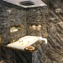 Luksusowa łazienka z wanną jacuzzi, wykończona ciemnymi płytkami imitującymi marmur, z złotymi akcentami na listwach i armaturze. Wnęki w ścianach z oświetleniem eksponują dekoracje.