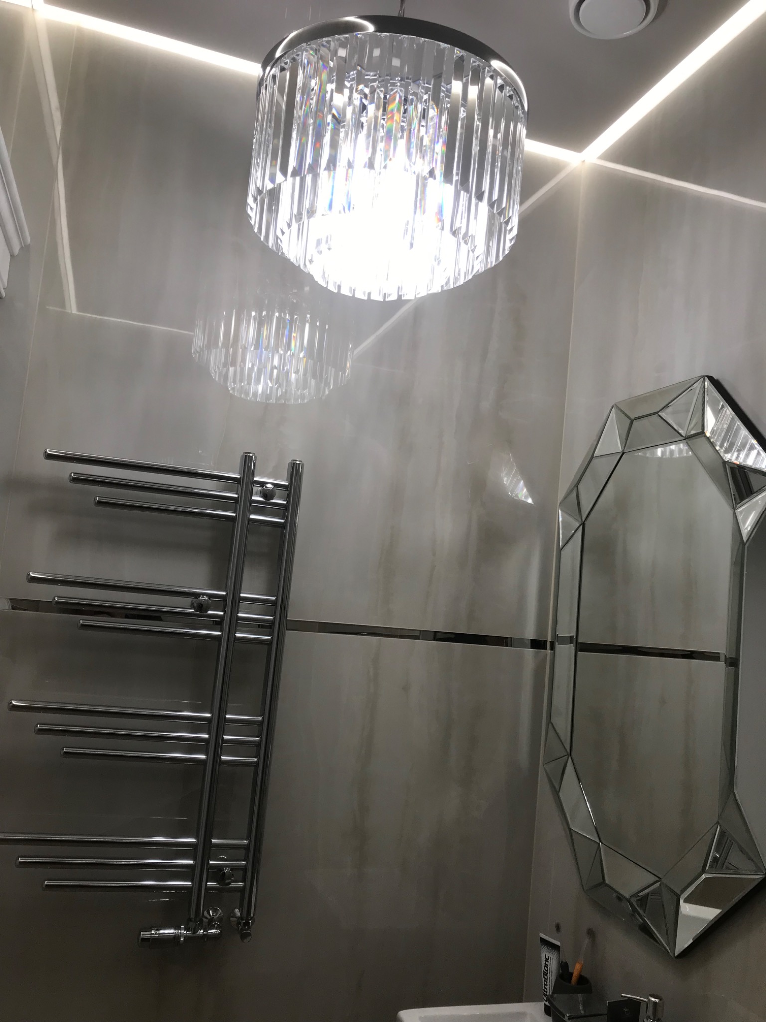 Nowoczesna łazienka z szarymi płytkami, kryształowym żyrandolem, grzejnikiem drabinkowym i lustrem w geometrycznej ramie.