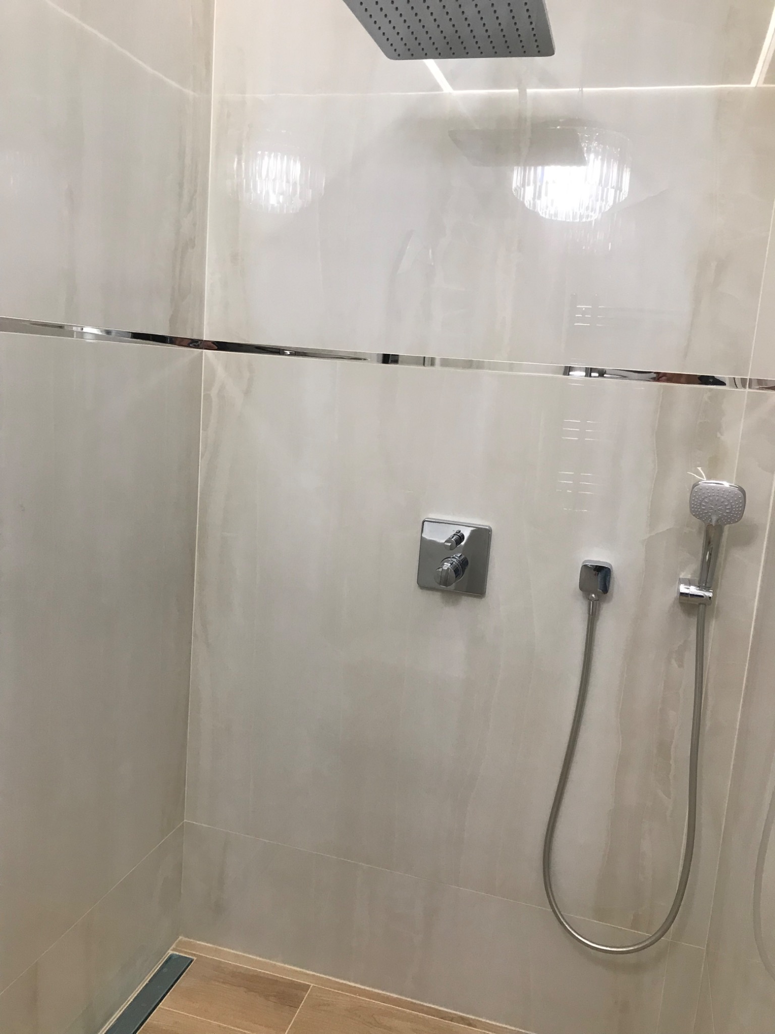 Wykończona kabina prysznicowa z beżowymi, marmurkowymi płytkami, chromowaną armaturą, deszczownicą i odpływem liniowym w podłodze z drewnopodobnych płytek.