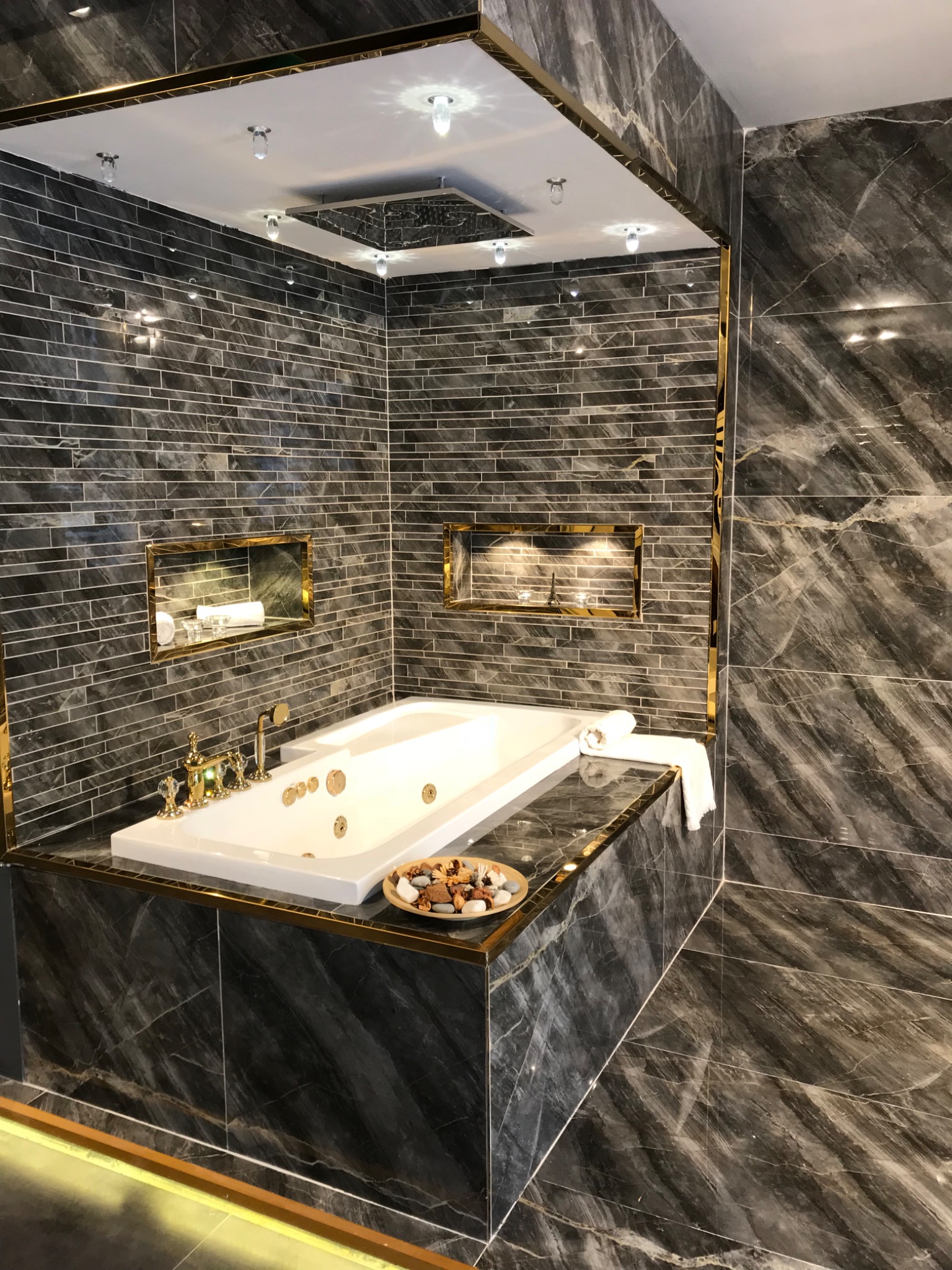 Luksusowa łazienka z wanną jacuzzi, wykończona ciemnymi płytkami imitującymi marmur, z złotymi akcentami na listwach i armaturze. Wnęki w ścianach z oświetleniem eksponują dekoracje.
