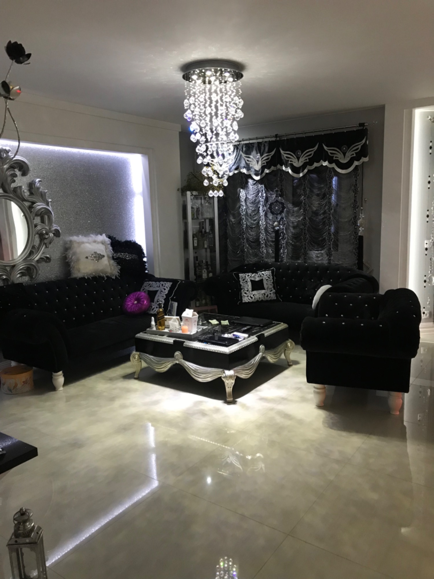 Elegancki salon w stylu glamour z czarnymi sofami obitymi aksamitem i kryształowym żyrandolem, błyszcząca podłoga odbija światło, w tle dekoracje okienne z motywem skrzydeł.