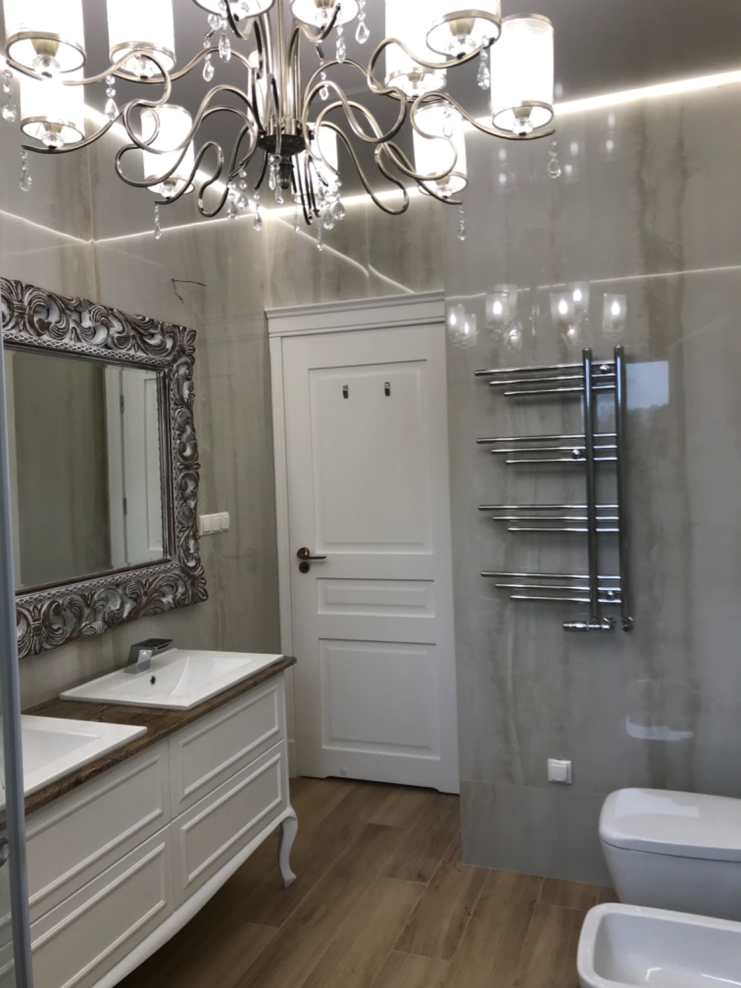 Elegancka łazienka z dwoma umywalkami na szafce z frezowanymi frontami, ozdobnym lustrem w ramie, chromowanym grzejnikiem drabinkowym i podłogą imitującą drewno, oświetlona dekoracyjnym żyrandolem.