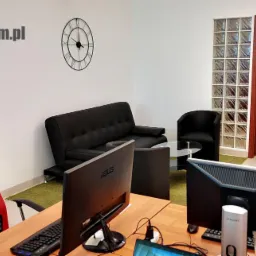 Nowoczesne biuro IT z dwoma stanowiskami komputerowymi, czerwonymi fotelami gamingowymi, czarną sofą i zegarem ściennym, logo firmy widoczne w rogu