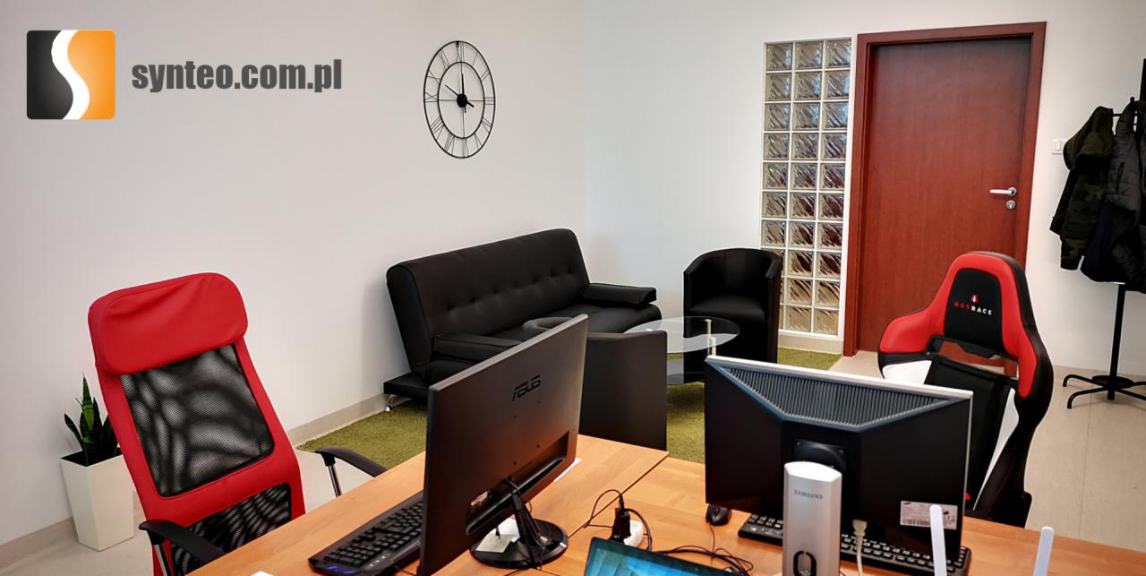 Nowoczesne biuro IT z dwoma stanowiskami komputerowymi, czerwonymi fotelami gamingowymi, czarną sofą i zegarem ściennym, logo firmy widoczne w rogu