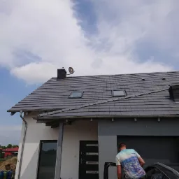 Antena satelitarna zamontowana na kominie nowego domu z szarą dachówką, widok z dołu, z fragmentem samochodu i osoby wychodzącej z niego.