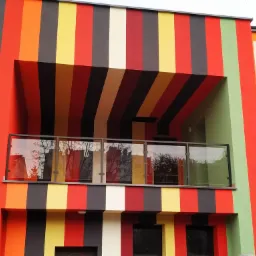 Kolorowa fasada budynku z balkonem zabezpieczonym szklaną balustradą, odbijającą drzewa i budynki.