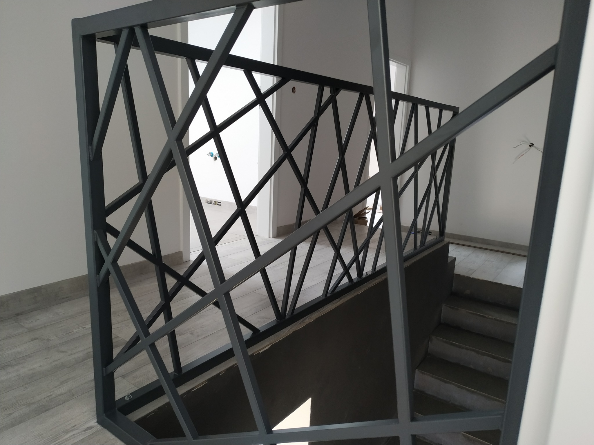 Nowoczesna balustrada schodowa z metalowych profili o geometrycznym wzorze rombów i trójkątów, szara kolorystyka, widok na schody i jasne wnętrze.