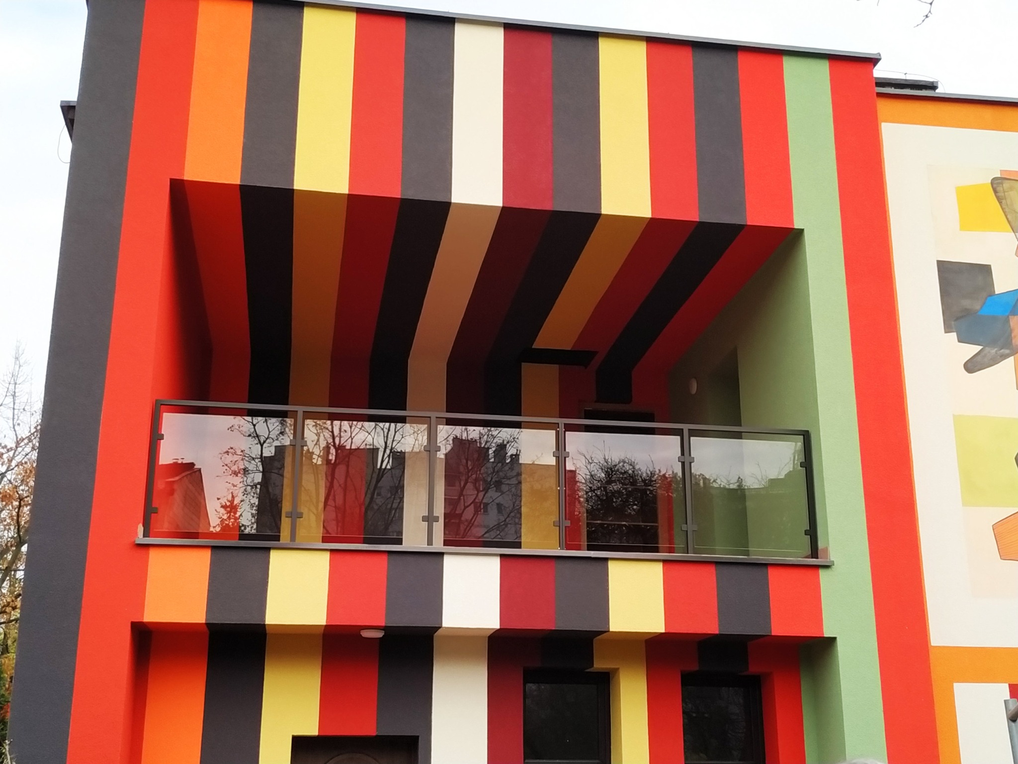 Kolorowa fasada budynku z balkonem zabezpieczonym szklaną balustradą, odbijającą drzewa i budynki.