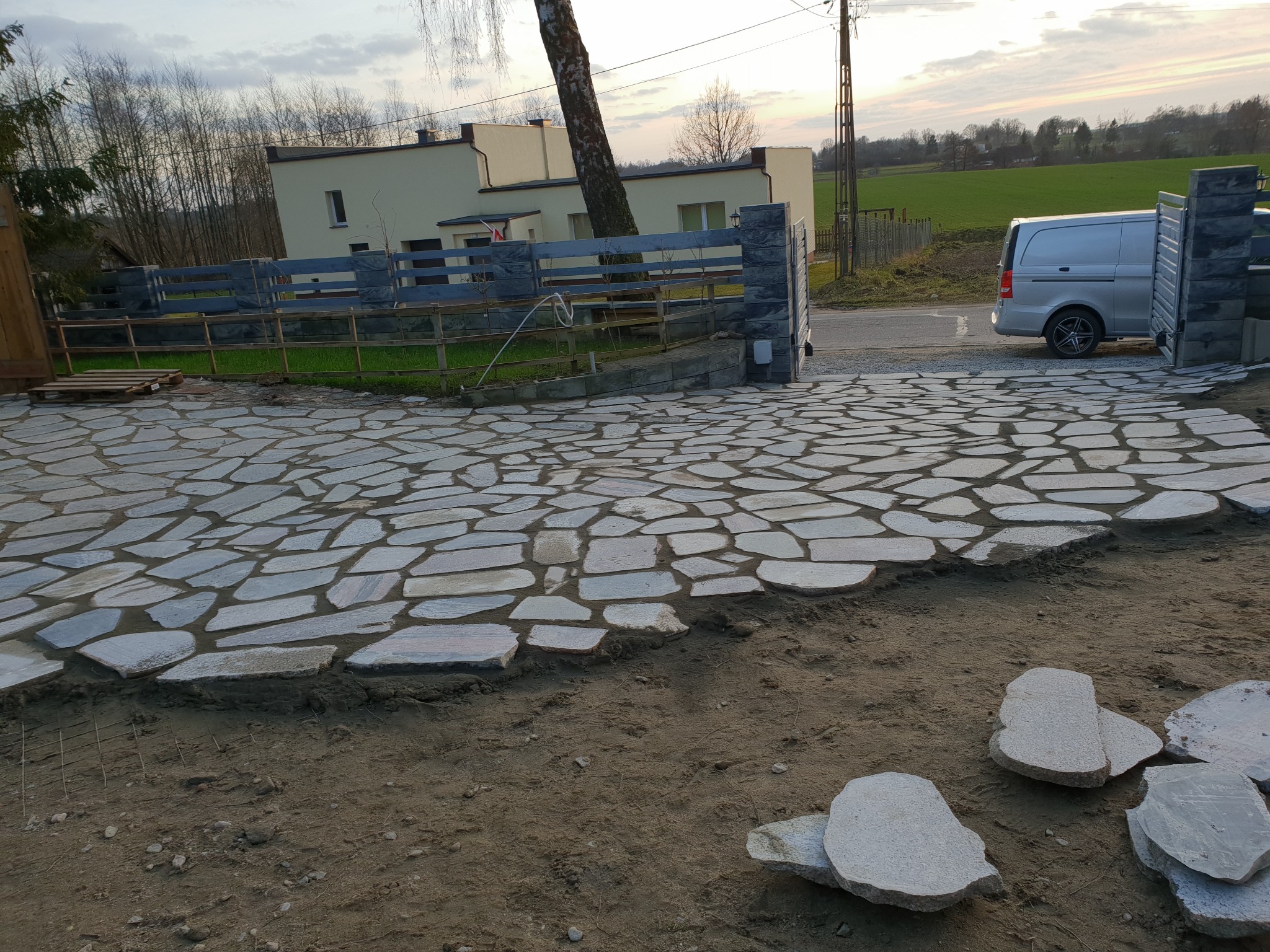 Układanie kamiennej nawierzchni na podjeździe, widoczny fragment ułożonej ścieżki z nieregularnych płyt, przygotowany teren i zaparkowany srebrny van.