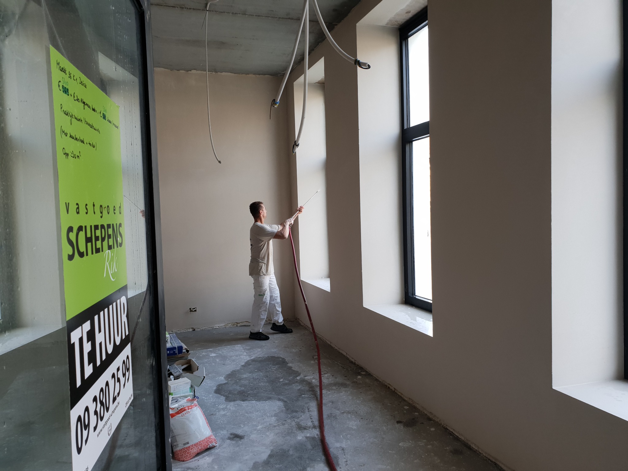 Malowanie ścian natryskowo w nowym budynku z dużymi oknami, widoczne wystające kable i plakat na szybie z ogłoszeniem o wynajmie.