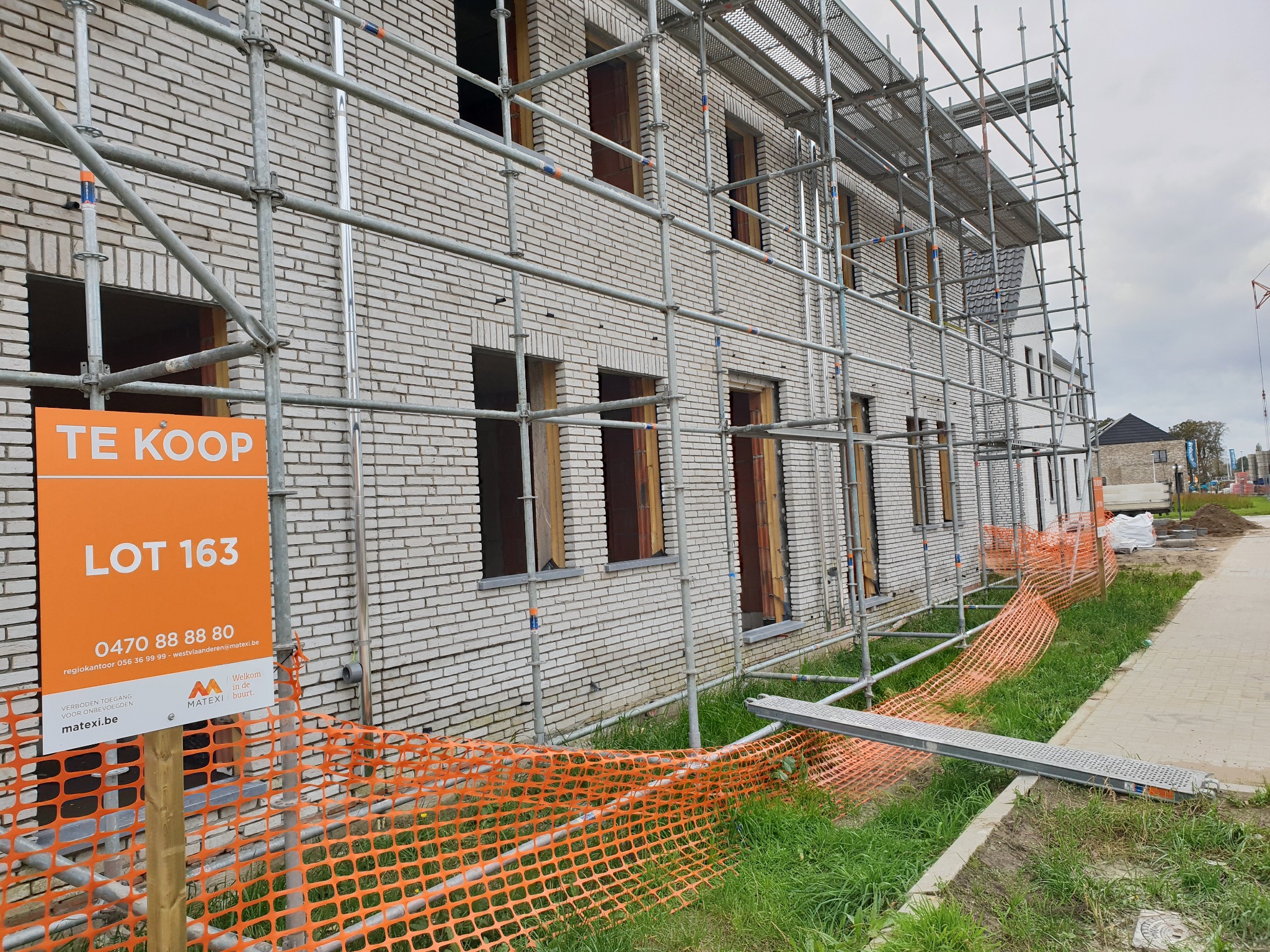 Budynek w trakcie budowy z białej cegły, otoczony rusztowaniem; pomarańczowa tablica informacyjna 'TE KOOP LOT 163' z numerem telefonu i logo firmy deweloperskiej.