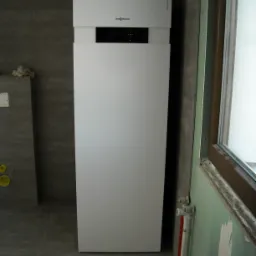 Biały, pionowy piec grzewczy Viessmann Vitocal 222 w szarej łazience z oknem. Widoczne rury instalacyjne i fragment ściany w kolorze zielonym.