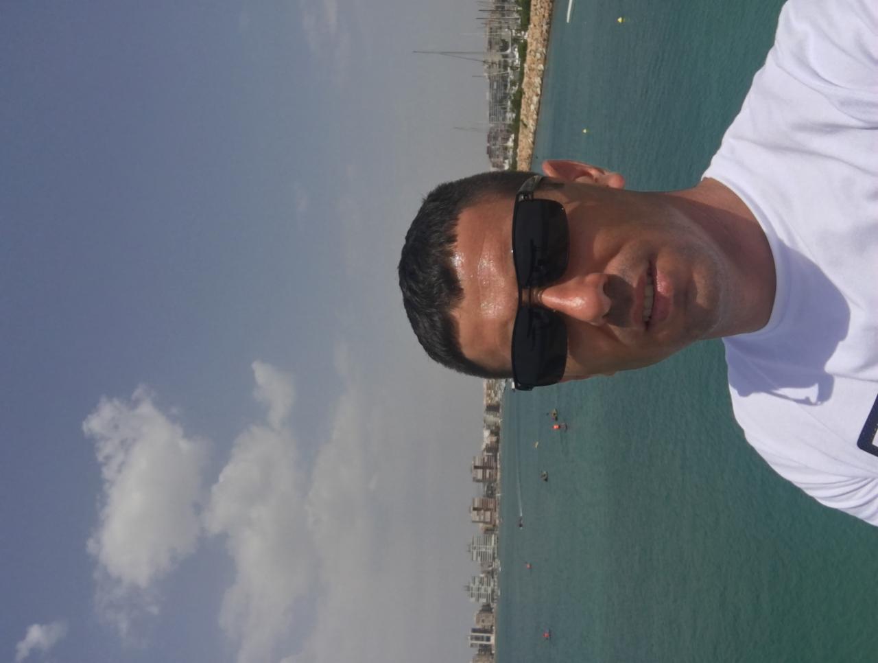 Mężczyzna w okularach przeciwsłonecznych pozuje na tle morza i zabudowy miejskiej, widoczny fragment białej koszulki.