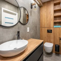 Nowoczesna łazienka z owalną umywalką nablatową, czarną armaturą, okrągłym lustrem w czarnej ramie, drewnianymi szafkami i szarymi płytkami.