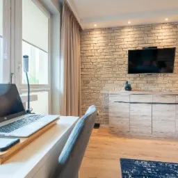 Jasne biuro domowe z ceglaną ścianą, biurkiem z laptopem, szarą tapicerowaną krzesłem i szafką RTV z telewizorem