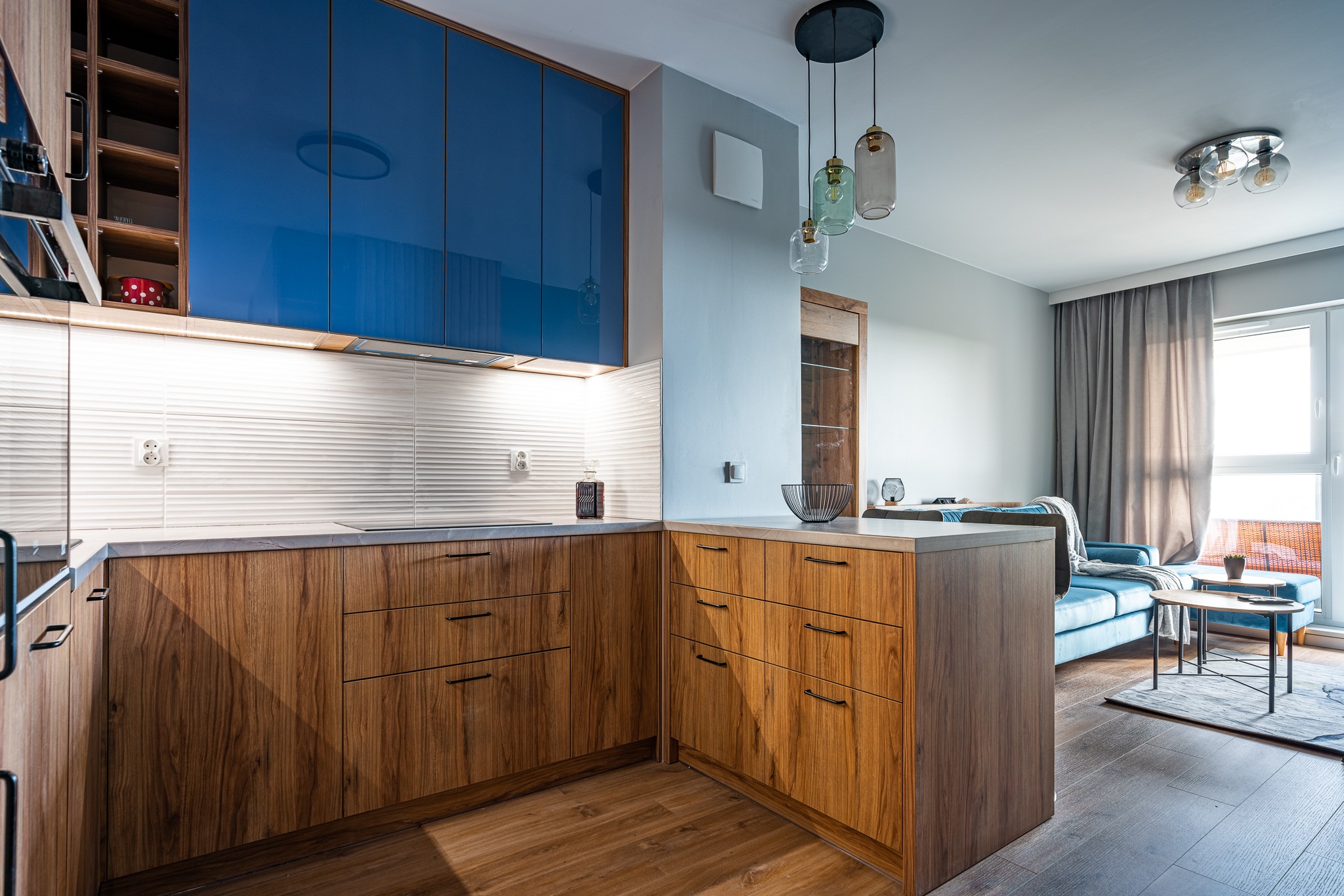 Nowoczesna kuchnia z drewnianymi szafkami dolnymi i błyszczącymi, niebieskimi szafkami górnymi, białymi płytkami ściennymi z teksturą fal, oraz salonem widocznym w tle z błękitną sofą i szarymi...