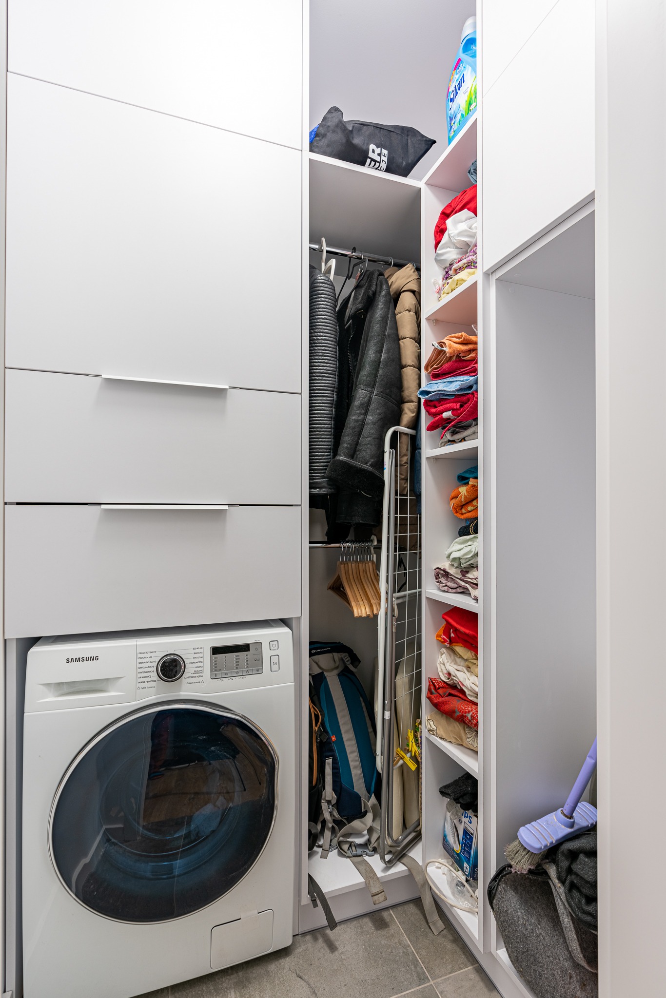 Wnęka z zabudową meblową: pralka pod szufladami, obok szafa z ubraniami, deską do prasowania, plecakiem i półkami z ręcznikami oraz środkami czystości.