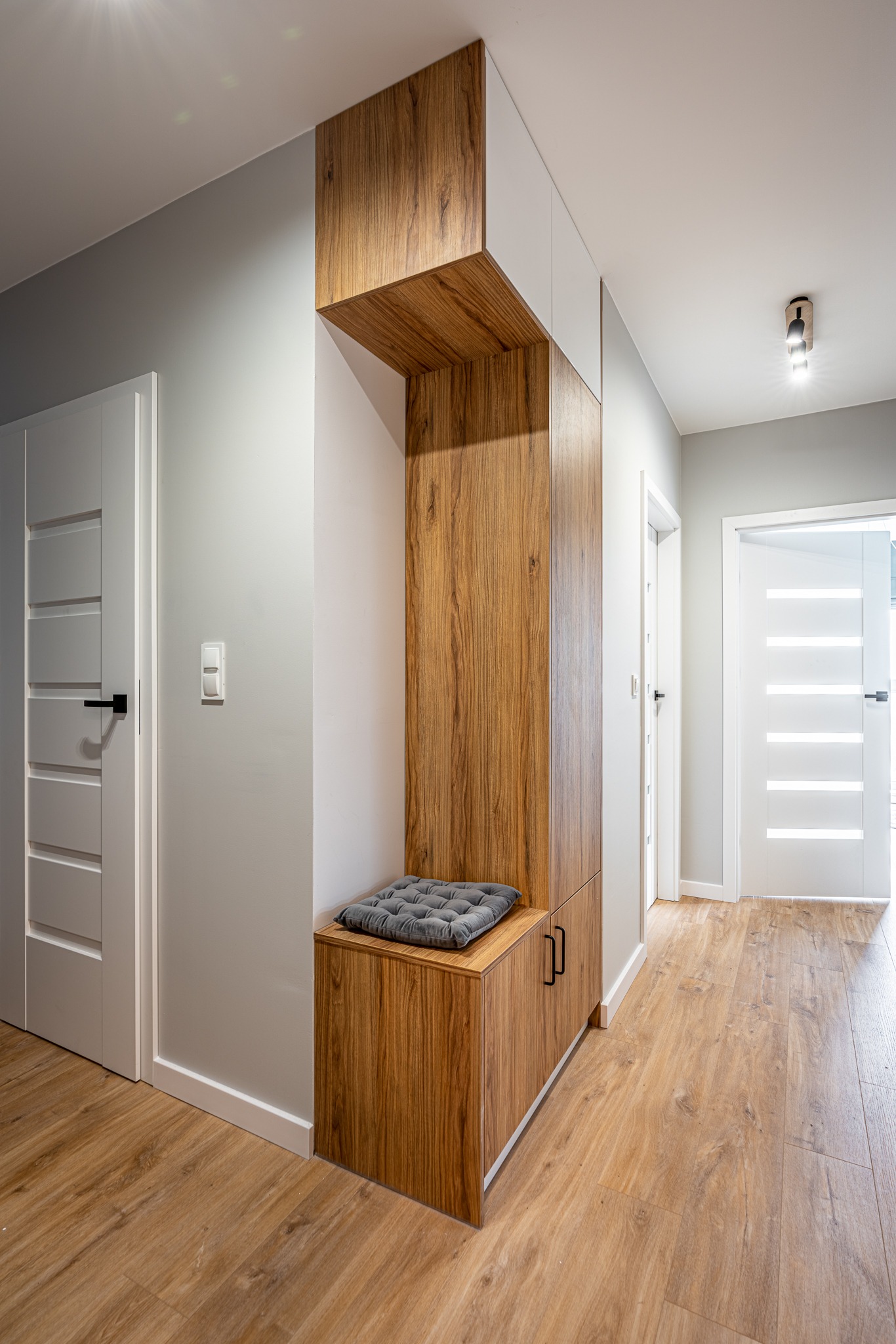 Nowoczesny przedpokój z drewnianą zabudową: szafa z siedziskiem i szarą poduszką, jasne ściany i podłoga imitująca drewno, białe drzwi z czarnymi klamkami.