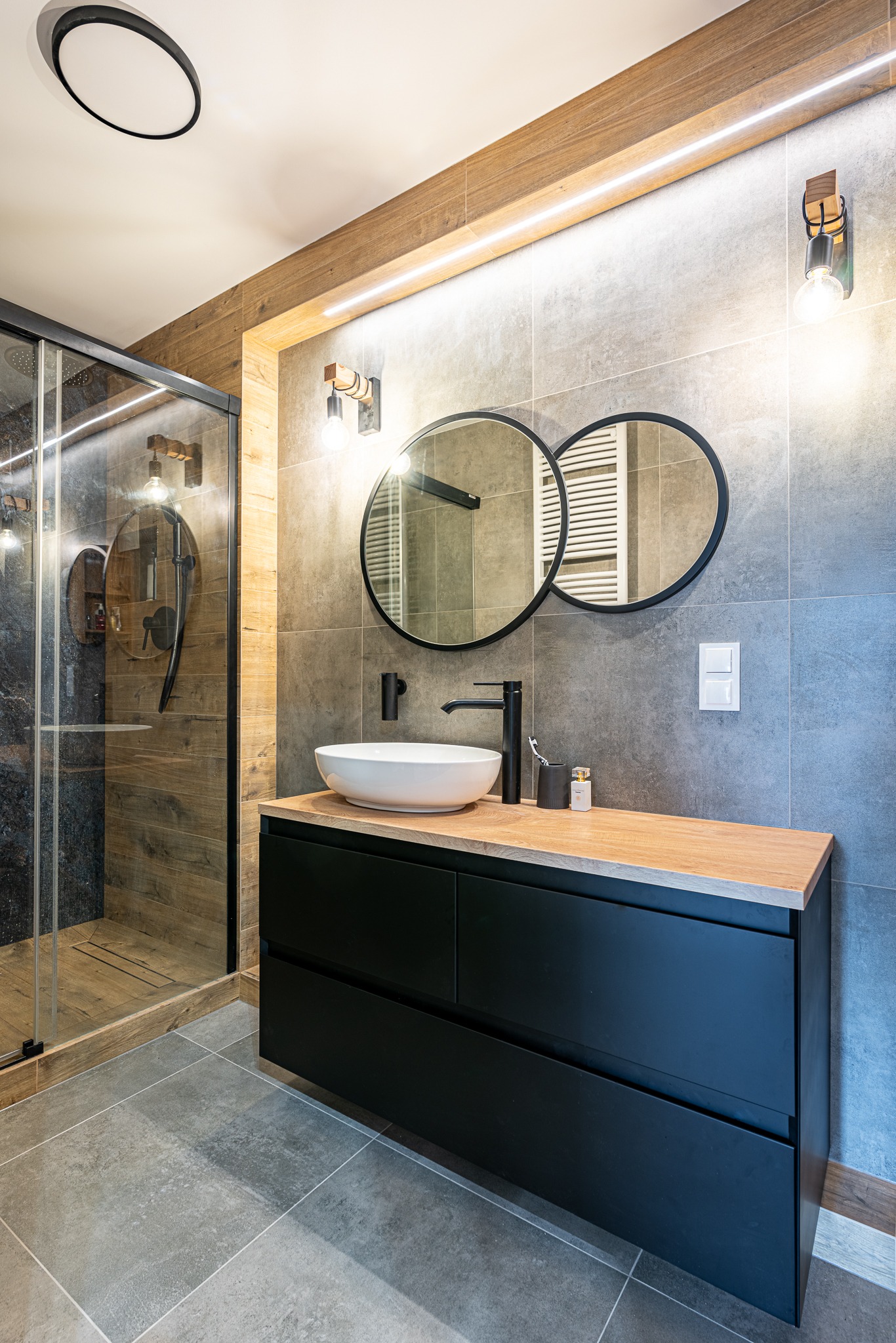 Nowoczesna łazienka z kabiną prysznicową typu walk-in, umywalką nablatową na czarnej szafce z drewnianym blatem, dwoma okrągłymi lustrami i szarą płytką imitującą kamień na ścianach.