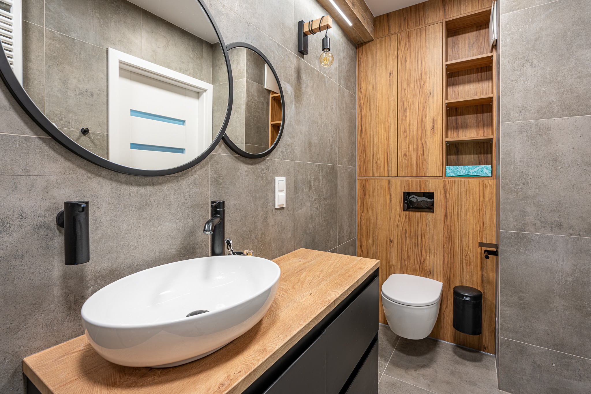 Nowoczesna łazienka z owalną umywalką nablatową, czarną armaturą, okrągłym lustrem w czarnej ramie, drewnianymi szafkami i szarymi płytkami.