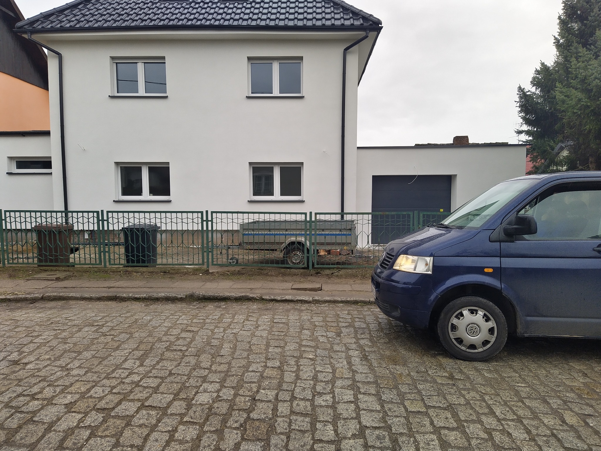 Biały dom jednorodzinny z nową elewacją, widoczny z poziomu ulicy wyłożonej kostką brukową, z zaparkowanym niebieskim vanem.