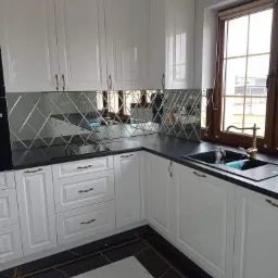 Kuchnia z białymi szafkami w stylu klasycznym, czarnym blatem i lustrzaną mozaiką w romby nad blatem, odbijającą okno w drewnianej ramie i złoty kran.