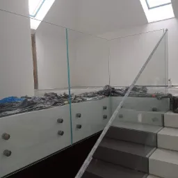 Szklana balustrada zamontowana na schodach w nowoczesnym wnętrzu, zabezpieczona folią ochronną podczas montażu, widoczne okna dachowe.