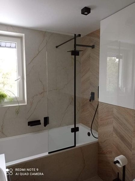 Nowoczesna łazienka z wanną i szklanym parawanem nawannowym z czarnymi elementami mocującymi, marmurowe i drewnopodobne płytki na ścianach, oświetlenie punktowe.