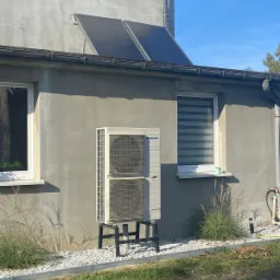 Zewnętrzna jednostka klimatyzacji Panasonic na czarnym stelażu przed szarą ścianą budynku z dwoma panelami słonecznymi na dachu, widoczne białe kamienie dekoracyjne i trawa.