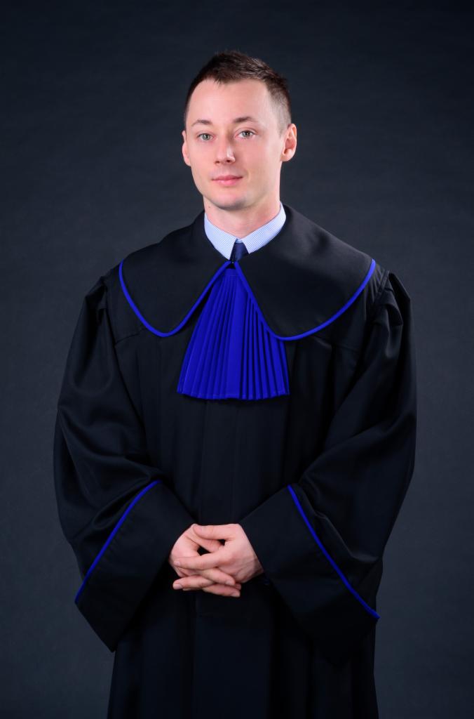 Portret młodego mężczyzny w todze prawniczej z niebieskimi akcentami, ręce splecione na wysokości pasa, na ciemnoszarym tle.