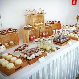Słodki stół z różnorodnymi deserami: babeczki z owocami, mini tartaletki, desery w słoiczkach, kokosowe kulki, drewniane skrzynki jako ekspozycja, dekoracja 'LOVE' w tle ściana z gaśnicą.