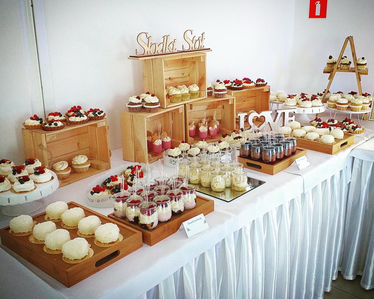 Słodki stół z różnorodnymi deserami: babeczki z owocami, mini tartaletki, desery w słoiczkach, kokosowe kulki, drewniane skrzynki jako ekspozycja, dekoracja 'LOVE' w tle ściana z gaśnicą.
