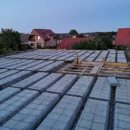 Wieniec stropowy z szalunkiem i zbrojeniem na budowie domu jednorodzinnego, widok z g&oacute;ry na tle zabudowy mieszkalnej o zmierzchu.