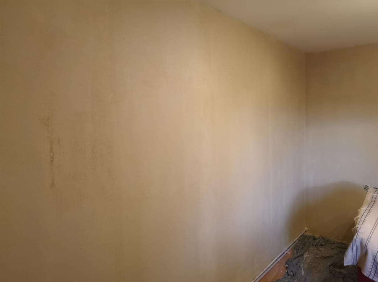 Świeżo otynkowana ściana w rogu pomieszczenia, widoczne niedoskonałości i zacieki, podłoga zabezpieczona folią malarską, przygotowana do dalszych prac wykończeniowych.