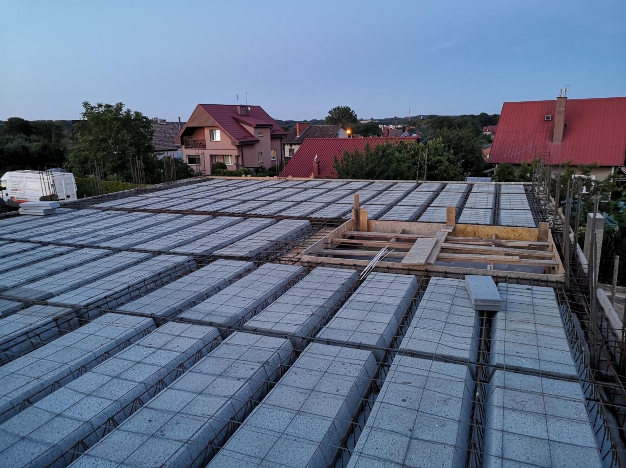 Wieniec stropowy z szalunkiem i zbrojeniem na budowie domu jednorodzinnego, widok z góry na tle zabudowy mieszkalnej o zmierzchu.