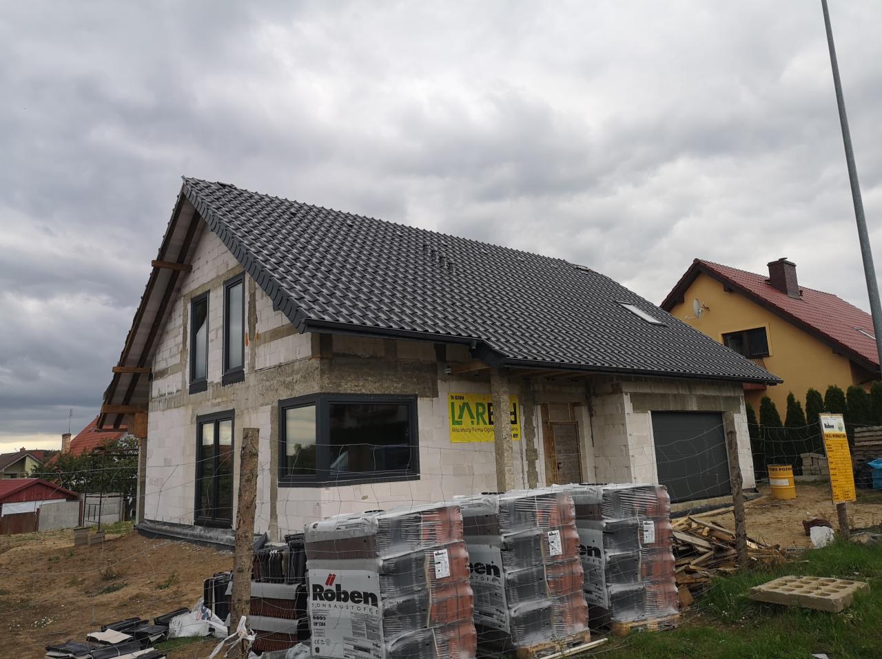 Niedokończony dom jednorodzinny z szarej cegły z czarną dachówką, czarnymi oknami i paletami cegieł Roben na pierwszym planie, w tle pochmurne niebo.