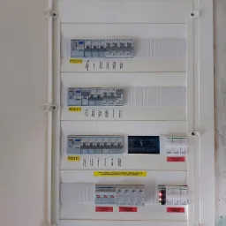 Otwarta skrzynka rozdzielcza z wyłącznikami różnicowoprądowymi i bezpiecznikami, widoczne oznaczenia obwodów oraz naklejka z danymi kontaktowymi firmy elektrycznej, surowa ściana w tle.