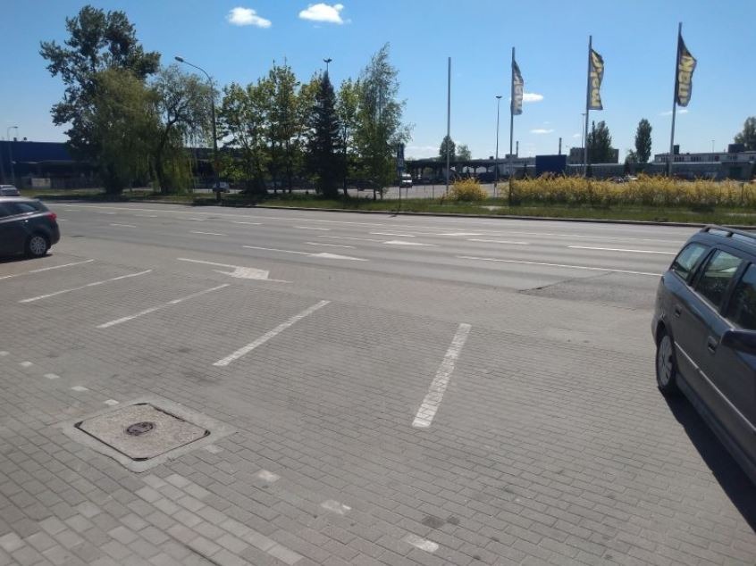 Parking z wyznaczonymi miejscami, samochody osobowe, w tle budynek z flagami reklamowymi w słoneczny dzień.