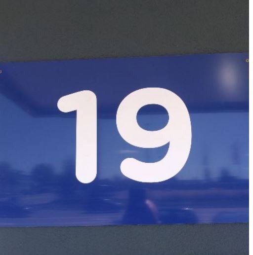 Niebieska tabliczka z białym numerem 19, przymocowana do ściany, widoczne odbicie krajobrazu.