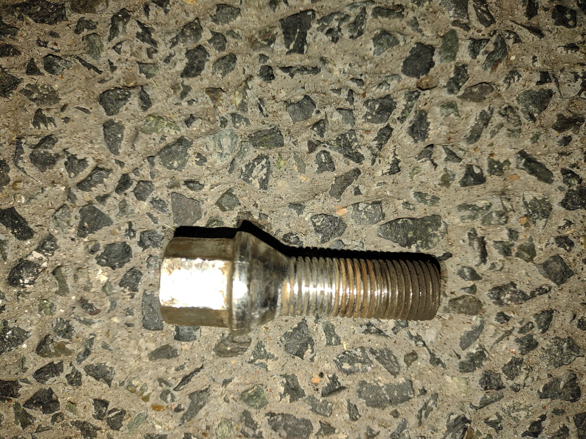 Śruba koła samochodowego z gwintem, leżąca na szorstkiej betonowej nawierzchni z widocznymi kamyczkami.