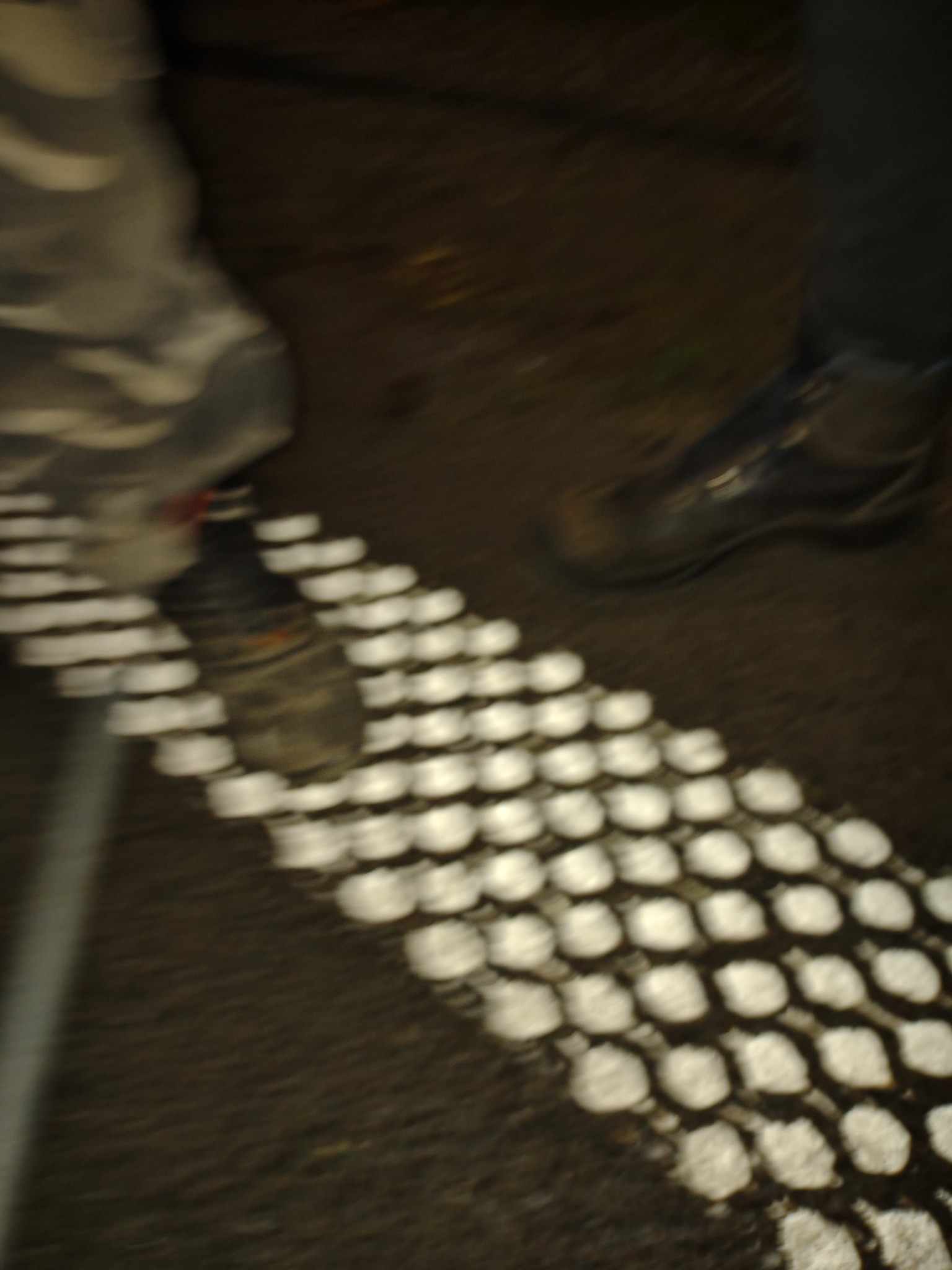 Niejasne zdjęcie fragmentu przejścia dla pieszych z widocznymi butami dwóch osób, jedna w wojskowych spodniach, druga w ciemnych spodniach.