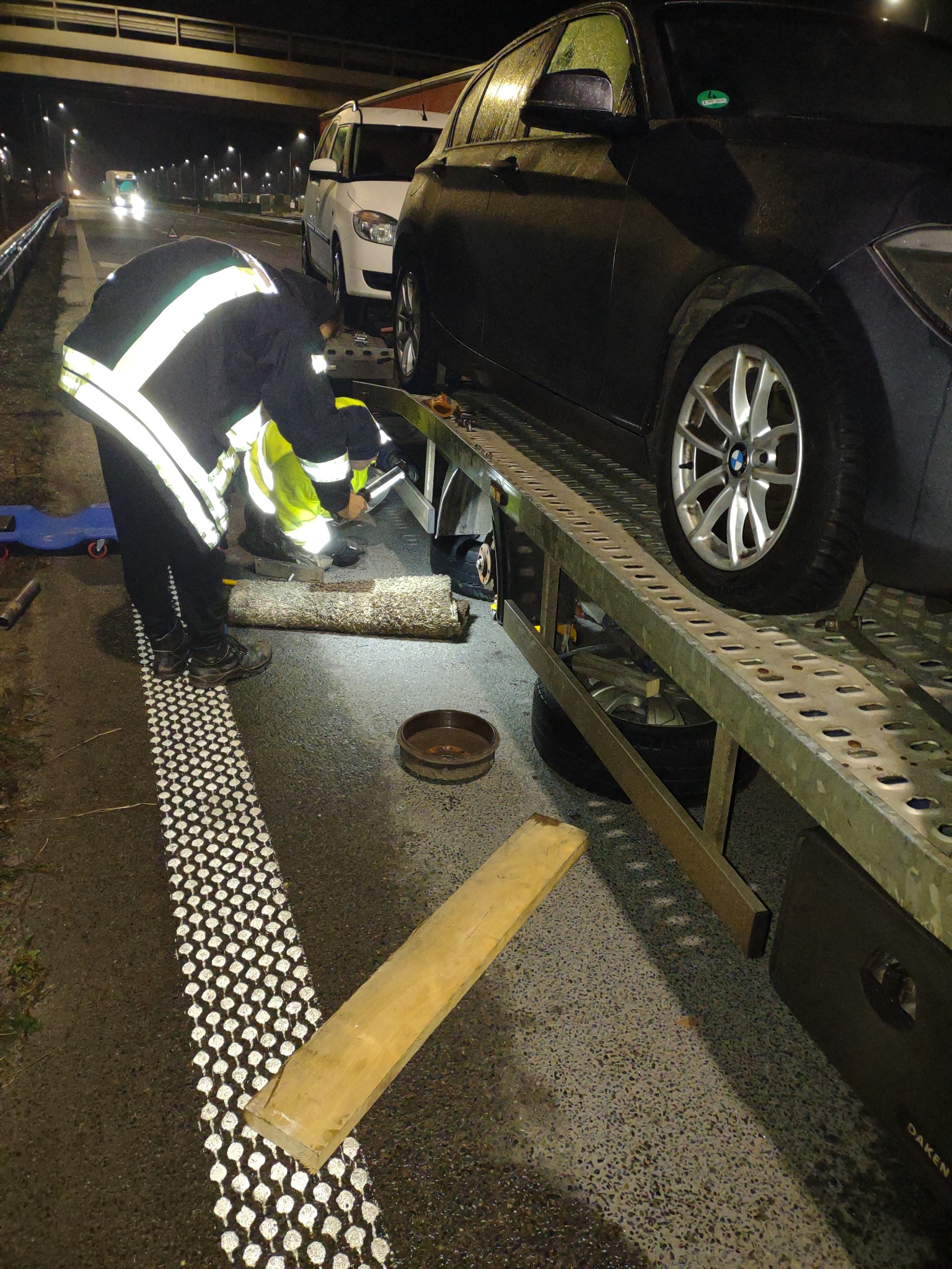 Czarny samochód BMW na lawecie w nocy, pracownik w odblaskowej kurtce przy kole, widoczne narzędzia i fragment drogi.