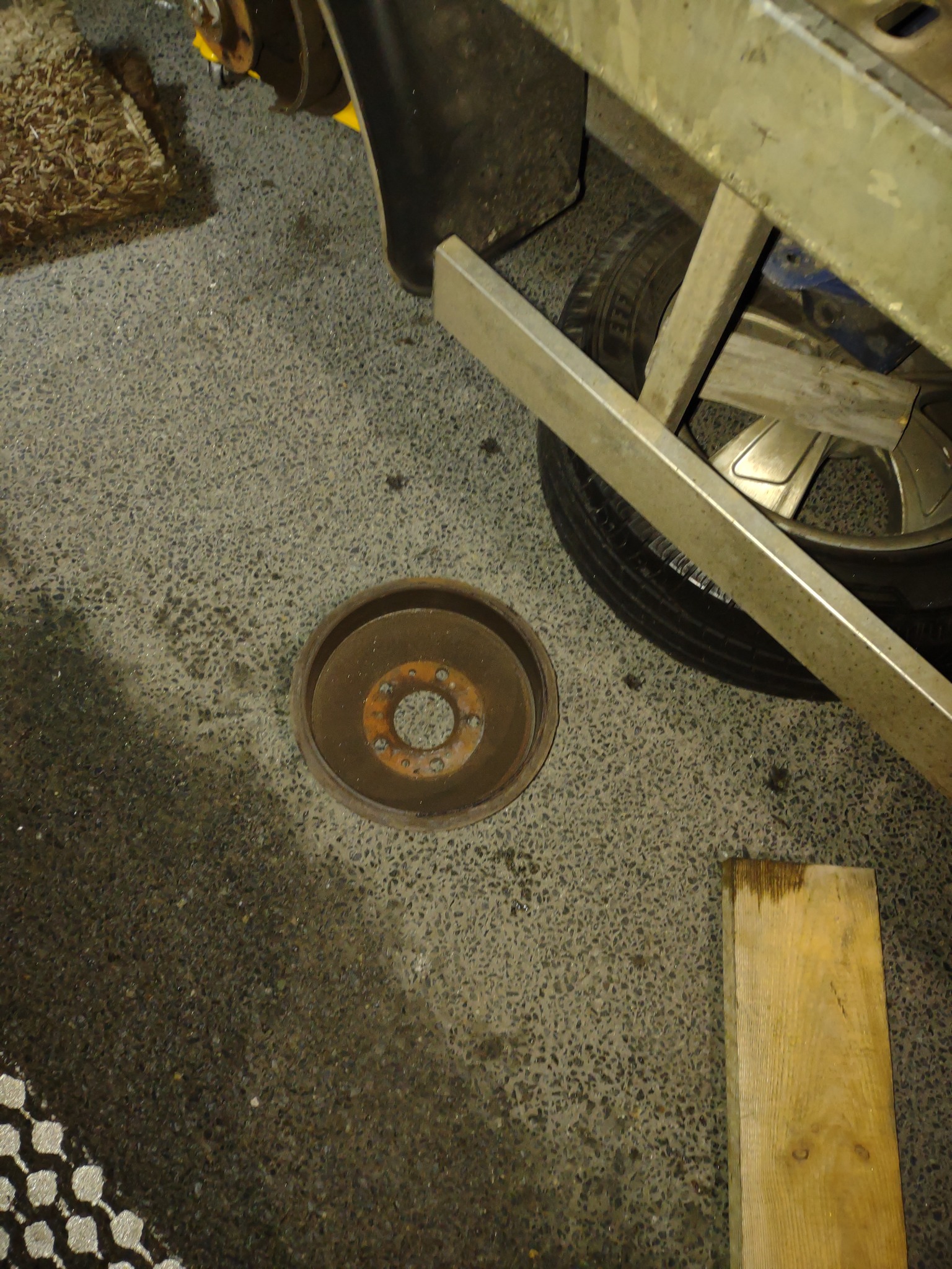 Zardzewiały bęben hamulcowy leżący na betonowej posadzce warsztatu, obok opony i metalowych elementów konstrukcyjnych.