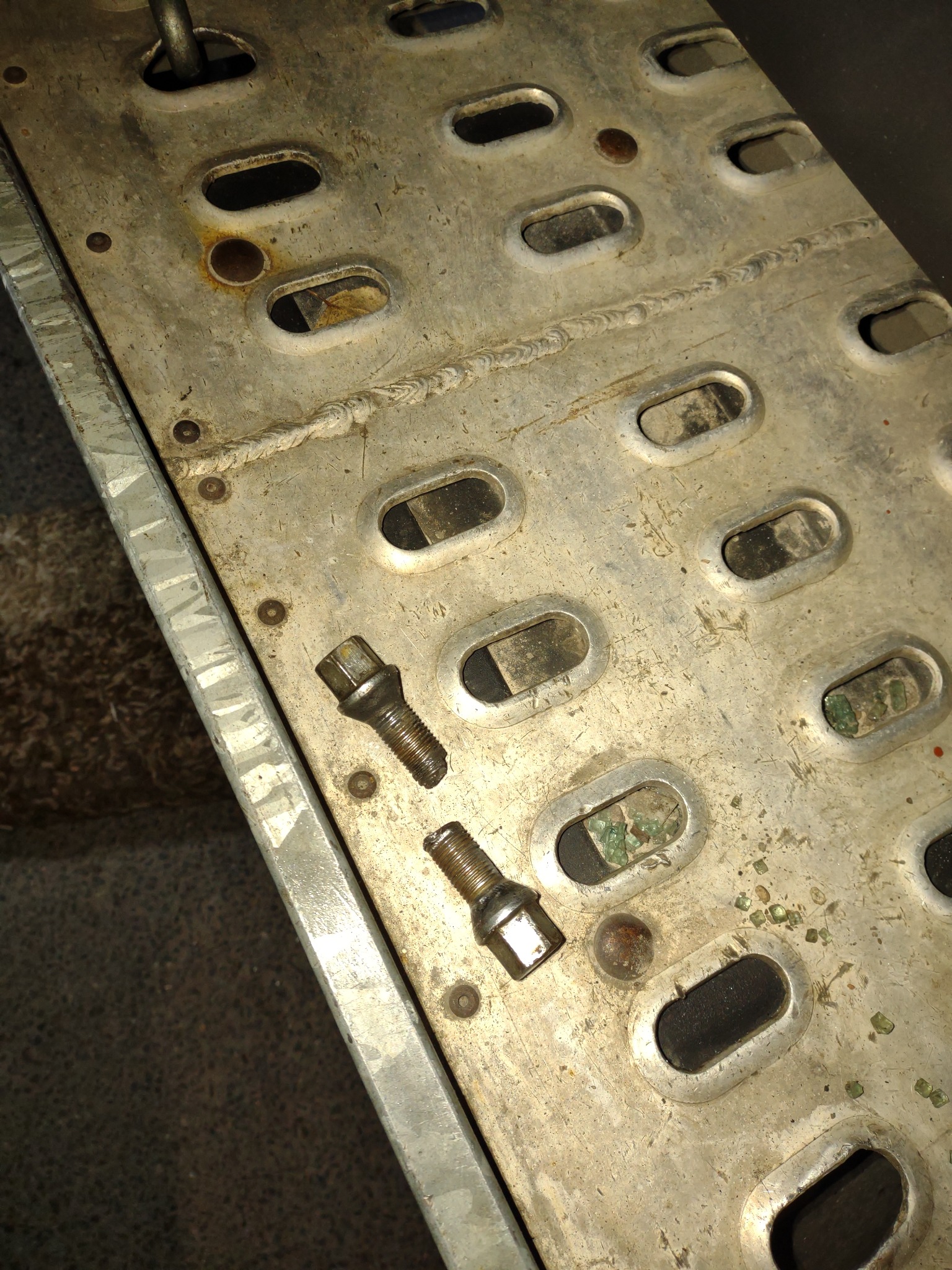 Metalowa rampa z owalnymi otworami, widoczne ślady użytkowania, rdza, dwa odkręcone śruby leżące na powierzchni.