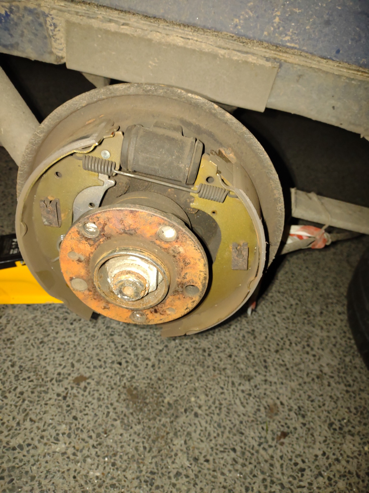 Zdjęcie z bliska przedstawia rozmontowany, skorodowany bęben hamulcowy pojazdu podczas naprawy, widoczny na podnośniku, z elementami mocującymi i sprężynami.
